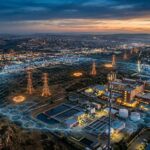 Aerial dusk view of a city with power pylons, water treatment tanks and a hexagonal digital shield glowing over the