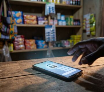 Hand reaching for a smartphone with a padlock on its screen, lying on a counter in a small shop with snacks on shelves